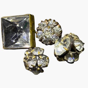 May include: Four pieces of jewelry with clear, faceted stones. One piece is a large, square-cut stone in a gold-toned setting. The other three pieces are clusters of smaller, round-cut stones in gold-toned settings.