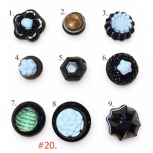May include: A collection of nine distinct black buttons labeled one through nine, featuring various shapes and center designs including white floral patterns, a gold glitter center, a green textured center, and a star-shaped white accent.