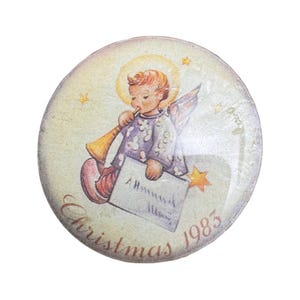 May include: A round decorative ornament featuring a vintage illustration of an angel with a golden halo playing a trumpet. The angel wears a purple patterned robe and holds a white card. The text reads Christmas 1983 in a cursive script.