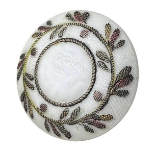 May include: A circular white button featuring a central embossed circle surrounded by a decorative border of embroidered metallic thread in shades of gold, silver, and copper, arranged in a leaf-like pattern across the textured white surface.