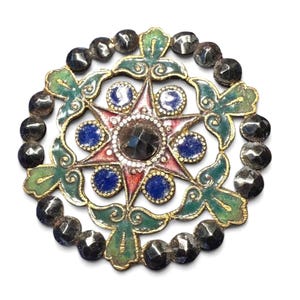 May include: A detailed, circular brooch with a starburst design. The brooch features a central dark gemstone surrounded by a ring of smaller white stones. The star has red and blue accents, with green and gold filigree details, and is encircled by dark, faceted beads.