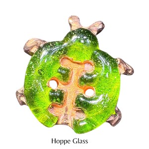 May include: A green glass button with a copper-colored design in the shape of a tree. The button has a sparkly finish and is labeled "Hoppe Glass".