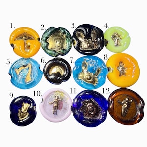 May include: A collection of twelve colorful, round glass buttons with gold-colored embossed designs. The buttons feature various designs, including figures, animals, and geometric shapes. The buttons are in shades of yellow, green, blue, and brown.