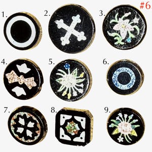 May include: A collection of nine small buttons with black backgrounds and gold-toned edges. The buttons feature various inlaid designs including white circles, crosses, floral patterns, and geometric shapes. One button is square, while the others are round.