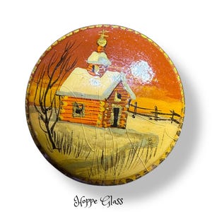 May include: A circular glass ornament featuring a hand-painted winter scene of a log cabin with a small steeple under a sunset sky. The artwork displays a bare tree, a wooden fence, and snow-covered ground. The text Hoppe Glass is printed below.