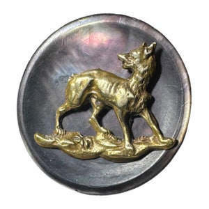 Può includere: Un bottone circolare con una base scura, iridescente e color carbone, caratterizzato da un rilievo metallico color oro raffigurante una volpe in piedi. La volpe è ritratta di profilo, rivolta a destra, con una texture del pelo dettagliata.