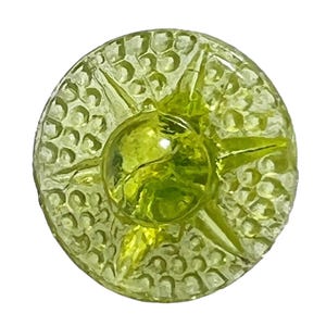 Può includere: Un bottone rotondo in vetro verde lime con un motivo a stella. Il bottone ha una superficie strutturata con piccoli rilievi e una cupola centrale arrotondata. Il bottone è posto su uno sfondo bianco.