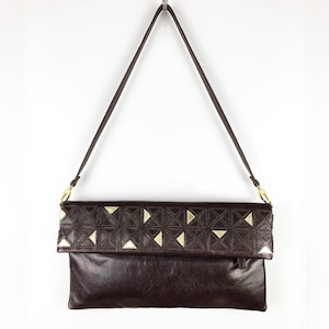 Könnte beinhalten: Dunkelbraune Leder-Umhängetasche mit langem Riemen. Die obere Klappe hat ein geometrisches Muster mit goldfarbenen Akzenten. Die Tasche hängt vor einem weißen Hintergrund.