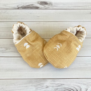 May include: A pair of baby booties in a mustard yellow color with a white geometric pattern of moons and crescents. The booties have a white fleece lining.