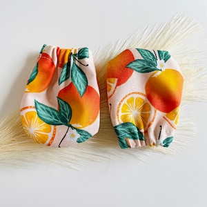 May include: A pair of orange and white fabric orange print hand warmers. The hand warmers are made of a soft, cotton fabric and feature a repeating pattern of oranges and leaves.