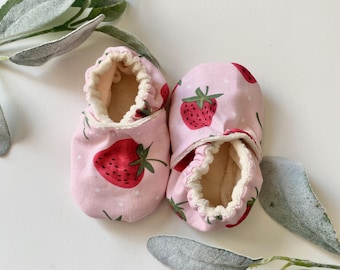 May include: A pair of baby booties with a pink background and a strawberry pattern. The booties have a soft, cream-colored interior and a ruffled edge. The strawberries are red with green leaves.