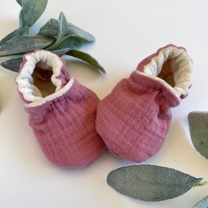 May include: A pair of soft, pink baby booties with white trim. The booties are made of a textured fabric and have a gathered elastic at the top.