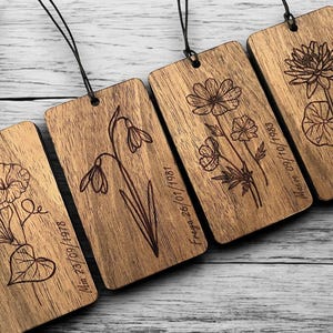 May include: Four wooden rectangular tags with engraved floral designs and dates. Each tag has a black string for hanging. The flowers include a morning glory, snowdrop, daisy-like flowers, and a water lily. The dates are engraved below the flowers.