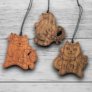 May include: Three wooden cat-shaped ornaments with black strings. Each cat represents a zodiac sign: Libra, Scorpio, and Virgo. The Libra cat holds scales, the Scorpio cat has a scorpion tail, and the Virgo cat wears a necklace and holds flowers.