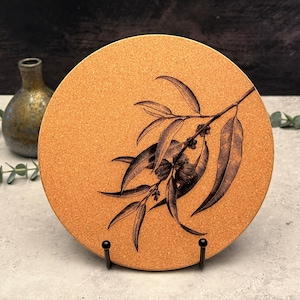May include: A circular cork coaster featuring a black illustration of eucalyptus leaves and flowers. The coaster is displayed on a black metal stand. A small, green and gold vase is in the background.