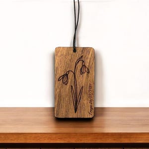 May include: A wooden tag with a black string, engraved with a snowdrop flower design. The tag has the name "Freya" and the date "26/01/1981" etched on it. The wood has a natural grain and is rectangular in shape.