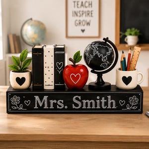 May include: A black rectangular wooden desk sign with the white text Mrs. Smith, flanked by a small potted plant, three decorative books, a red apple ornament, a black globe on a stand, and a white mug containing pencils and a wooden ruler.
