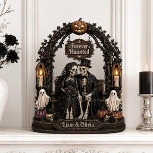 Può includere: Una statuetta decorativa di Halloween con due scheletri in abiti nuziali seduti su una panchina sotto un traliccio ad arco. L'arco mostra una zucca, il testo Forever Haunted e due fantasmi. La base reca l'iscrizione Liam & Olivia.