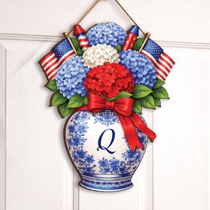 Könnte beinhalten: Patriotischer Türhänger mit einer Vase voller blauer, weißer und roter Blumen, amerikanischen Flaggen und Feuerwerk. Die Vase hat ein blau-weißes Blumenmuster und eine rote Schleife. Der Buchstabe "Q" befindet sich auf der Vase.