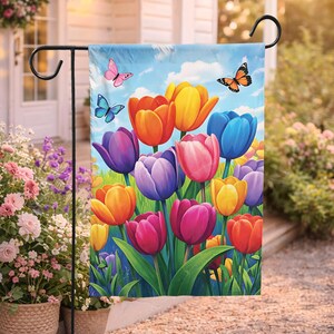 Può includere: Una colorata bandiera da giardino con una vivace illustrazione di tulipani nei toni del rosa, arancione, giallo e viola. Le farfalle svolazzano su uno sfondo di cielo blu. La bandiera è esposta su un supporto in metallo nero, ideale per l'arredamento esterno.