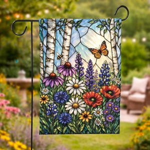 May include: A garden flag featuring a stained-glass design with birch trees, colorful flowers, and butterflies. The flowers include daisies, poppies, and purple coneflowers. A bee and an orange butterfly are also visible. The flag is displayed outdoors.