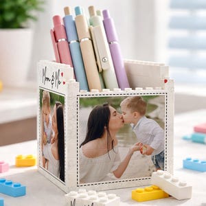 Może przedstawiać: Biały pojemnik na długopisy w stylu klocków z fotografią matki i dziecka oraz napisem "Mom & Me". Pojemnik zawiera kilka długopisów w pastelowych kolorach. Kolorowe klocki do zabawy są rozrzucone.