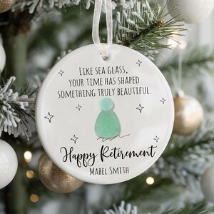 May include: A white ceramic Christmas ornament with a ribbon for hanging. The ornament features the text "LIKE SEA GLASS, YOUR TIME HAS SHAPED SOMETHING TRULY BEAUTIFUL." and "Happy Retirement" with a name below. The ornament is decorated with a sea glass design and star shapes.
