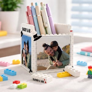 Può includere: Un portapenne bianco a forma di blocco con una foto di un padre e un bambino e la scritta "DADDY & ME". Il portapenne contiene diverse penne dai colori pastello. Blocchi giocattolo colorati e un personaggio Lego sono sparsi intorno al portapenne.
