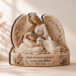 May include: A beige angel figurine with a mother and child design. The angel has large wings and a halo. The child is resting on a pillow. The base has the text "I HAVE AN ANGEL IN HEAVEN I CALL HER Mom 1965-2026". Pink flowers are on the base.