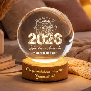 Może przedstawiać: Przezroczysta szklana kula z okazji ukończenia szkoły z napisem "Class of 2026", czapką absolwenta i dyplomem. Kula spoczywa na drewnianej podstawie z napisem "Congratulations on your Graduation!". Kula jest podświetlona od wewnątrz.