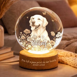 May include: A clear glass sphere with a detailed engraving of a dog, surrounded by flowers and butterflies, sits atop a wooden base. The base has the text "You left a paw print on our hearts 2020-2025" and the dog's name "Bailey".