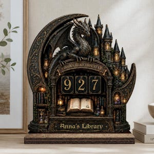 Könnte beinhalten: Eine dunkel gehaltene, halbmondförmige Dekofigur mit einem Drachen, der auf einer Burg thront. Das Objekt zeigt mittig die Zahlen 927, ein aufgeschlagenes Buch sowie den Schriftzug "BOOKS READ THIS YEAR" und "Anna's Library".