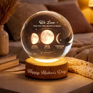 May include: A clear crystal ball with a wooden base, illuminated from within. The sphere features phases of the moon and the text "We Love You To The Moon & Back." Names and dates are inscribed below the moon phases. The base reads "Happy Mother's Day."