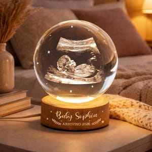 Op de afbeelding: Een gloeiende kristallen bol lamp met een echo-afbeelding van een baby. De lamp staat op een houten voet met de inscriptie "Baby Sophia" en het jaar "ARRIVING 2026". De lamp straalt een warm, zacht licht uit.