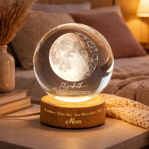 Op de afbeelding: Een heldere glazen bol met een gedetailleerd maan- en sterrenkaartontwerp, rustend op een houten voet. De voet is gegraveerd met de tekst "Under This Sky You Became My Mom". De bol wordt van binnenuit verlicht, waardoor een warme gloed ontstaat.