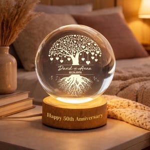 May include: A clear glass sphere with an engraved tree design, hearts, and the names "David & Anna" with the date "10.24.1976". The sphere sits on a wooden base with "Happy 50th Anniversary" engraved. The base is illuminated.