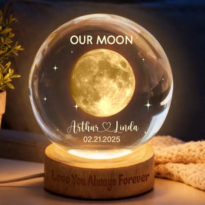 Könnte beinhalten: Eine klare Glaskugel mit einem leuchtenden Mond im Inneren, auf einem Holzsockel. Der Text "OUR MOON", die Namen "Arthur & Linda" und das Datum "02.21.2025" sind zu sehen. Auf dem Sockel steht "Love You Always Forever".
