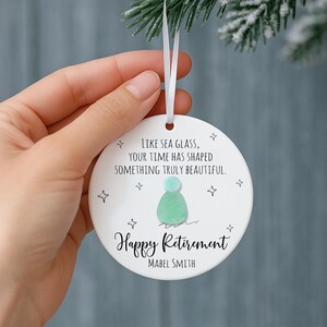 May include: White ceramic ornament with the text "LIKE SEA GLASS, YOUR TIME HAS SHAPED SOMETHING TRULY BEAUTIFUL." and "Happy Retirement" with a sea glass design. Includes the name "MABEL SMITH".