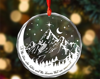 May include: A clear, round Christmas ornament with a mountain scene and the text "To the stars who listen and the dreams that are answered." The design includes a mountain range, stars, a crescent moon, and a star at the peak. It hangs from a red ribbon.