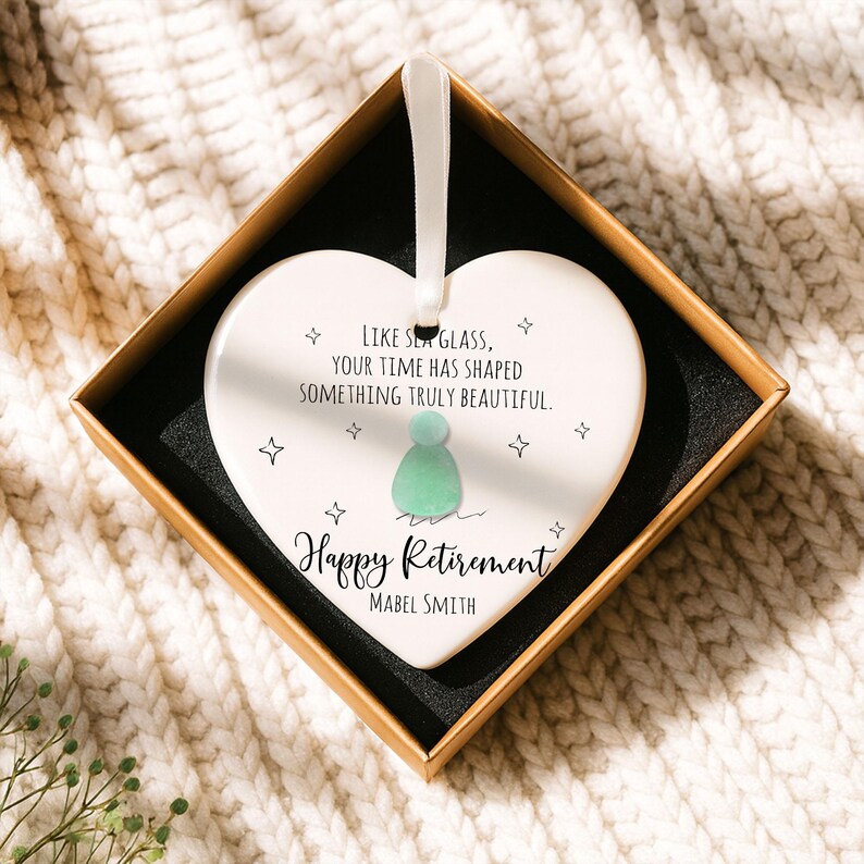 May include: A white heart-shaped ornament with a ribbon, inside a gift box. The ornament has the text "LIKE SEA GLASS, YOUR TIME HAS SHAPED SOMETHING TRULY BEAUTIFUL" and "Happy Retirement Mabel Smith". It features a green figure and star designs.