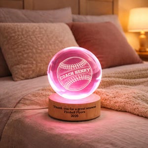 May include: A glowing pink baseball-themed night light. The clear glass orb features a baseball design with the text "COACH HENRY." The wooden base is engraved with "Thank you for a great season Fireball Flyers 2026."