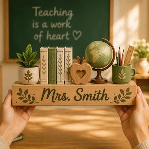 May include: A rectangular wooden desk organizer featuring the name Mrs. Smith engraved on the front. It holds a small potted plant, three miniature books, a wooden apple cutout, a small globe, and a green mug containing pencils and a ruler.