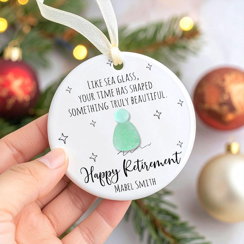 May include: A white ceramic ornament with a cream-colored ribbon. The ornament features the text "LIKE SEA GLASS, YOUR TIME HAS SHAPED SOMETHING TRULY BEAUTIFUL." and "Happy Retirement." A sea glass design is in the center. The name "MABEL SMITH" is below.