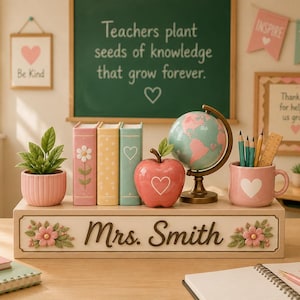 May include: A wooden desk organizer featuring the name Mrs. Smith in script, decorated with pink flowers. It holds a small pink potted plant, three pastel books, a red apple ornament with a heart, a small globe, and a pink mug with pencils.