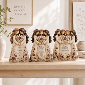 Pode incluir: Três estatuetas decorativas representando grupos de duas, três e quatro mulheres com cabelos longos, coroas de flores e vestidos cor de creme. Cada estatueta apresenta um coração central com detalhes florais e texto, incluindo You're the Sister I Got to Choose.
