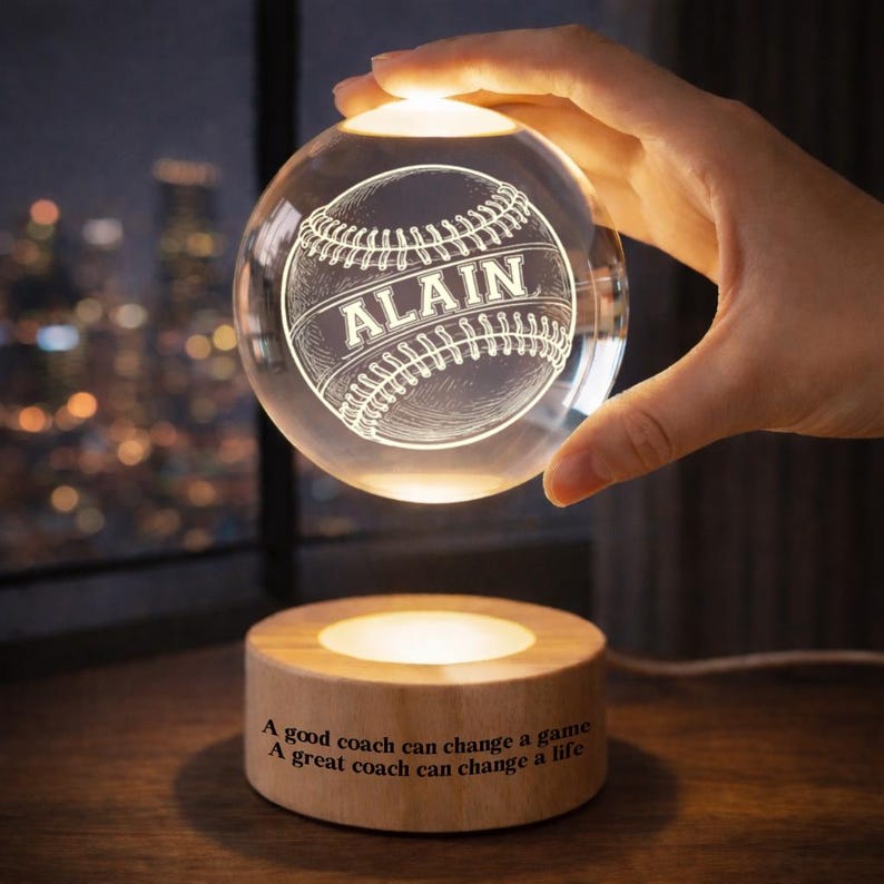 May include: A clear crystal baseball with the name "ALAIN" etched on it, illuminated from within. The baseball is held above a wooden base with the text "A good coach can change a game. A great coach can change a life."