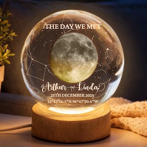 Może przedstawiać: Spersonalizowana kryształowa kula z motywem księżyca. Tekst "THE DAY WE MET", imiona "Arthur & Linda", data i współrzędne są wygrawerowane. Spoczywa na drewnianej podstawie, oświetlona ciepłym blaskiem.