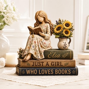 Könnte beinhalten: Eine Figur einer Person mit langem, welligem Haar in einem geblümten Kleid, die ein offenes Buch liest und auf einem Stapel aus drei Büchern sitzt. Die Bücher sind mit Elsa, Just a Girl und Who Loves Books beschriftet. Ein kleiner Krug mit Sonnenblumen steht daneben.