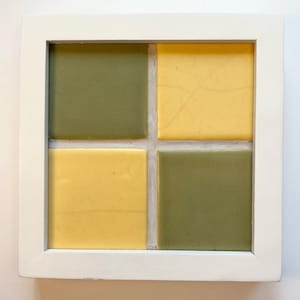 May include: A square, framed artwork featuring four square tiles. Two tiles are olive green, and two are yellow. The tiles are arranged in a 2x2 grid within a white wooden frame. The artwork has a minimalist, geometric design.
