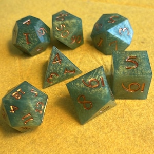 Puede incluir: Conjunto de siete dados verde azulado con números de color cobre. Los dados son de diferentes formas y tamaños.